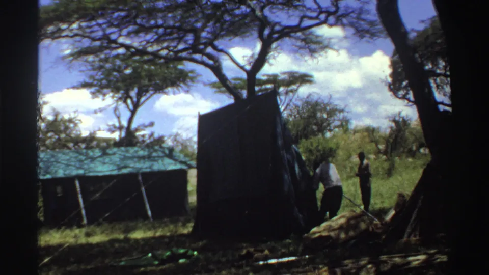 1969 Nigeria 8mm frame showing men beside a large dark tent under trees near a small green-roofed structure.