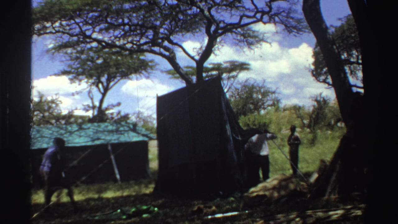 1969 Nigeria 8mm frame with a man moving across the shaded foreground as others adjust a dark camp tent.