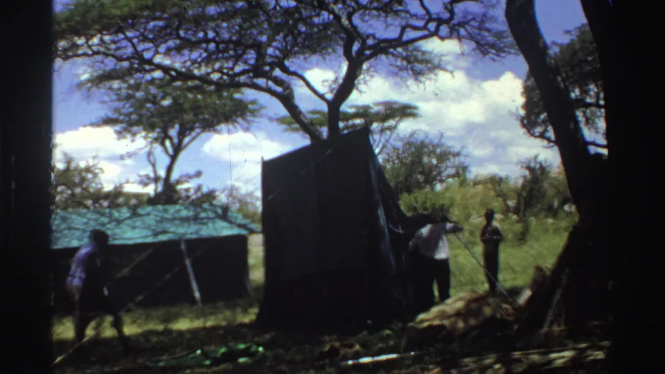 1969 Nigeria 8mm frame with a man moving across the shaded foreground as others adjust a dark camp tent.
