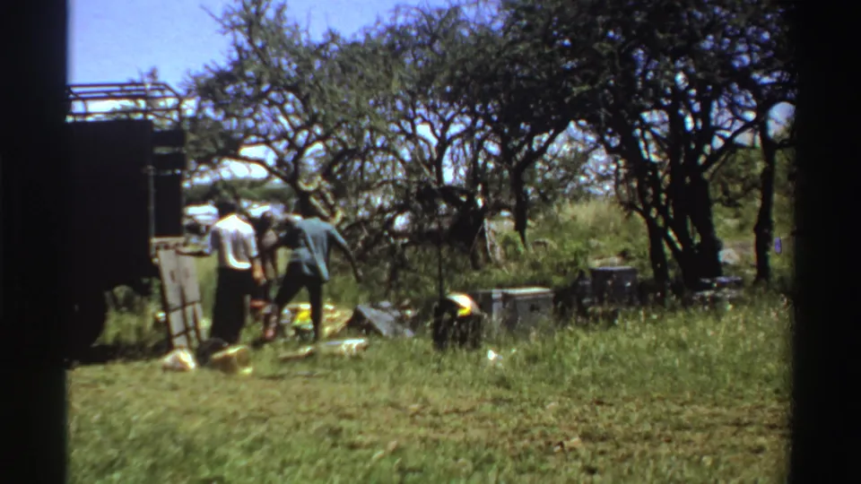 1969 Nigeria 8mm frame showing men beside a truck with scattered supplies in a grassy, tree-dotted outdoor area.