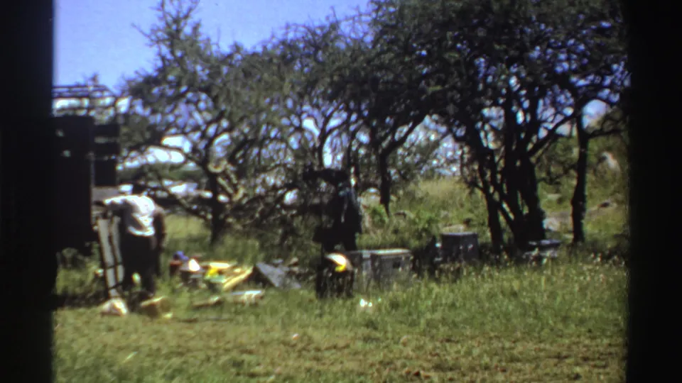1969 Nigeria home-movie frame showing a man by a truck and supplies spread across grass under trees.