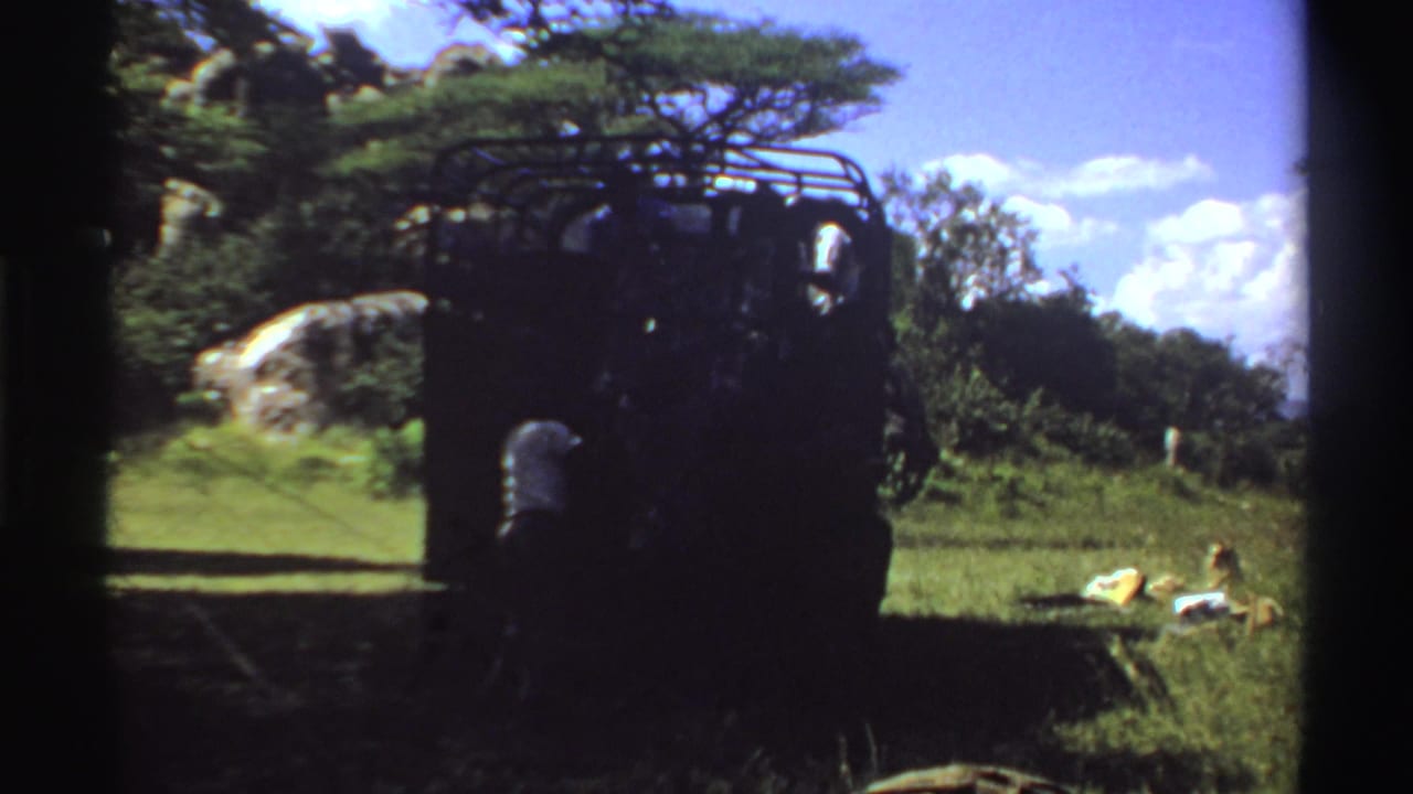 1969 Nigeria home-movie frame of a dark truck partly in shadow, with a nearby figure or object and supplies lying in the grass.