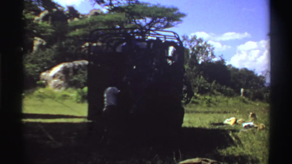 1969 Nigeria home-movie frame of a dark truck partly in shadow, with a nearby figure or object and supplies lying in the grass.