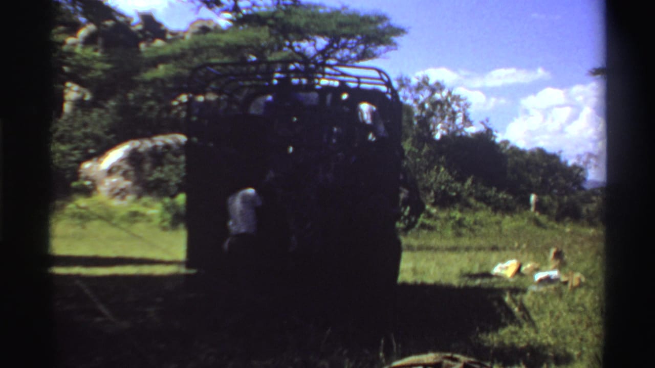 1969 Nigeria frame of a roof-rack truck in shade, with grassy terrain, trees, a rock outcrop, supplies, and distant people nearby.