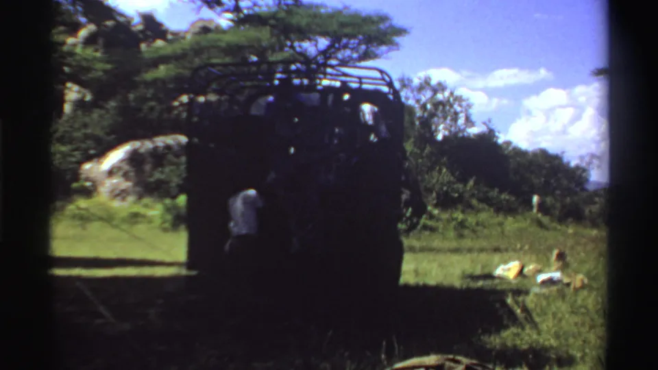 1969 Nigeria frame of a roof-rack truck in shade, with grassy terrain, trees, a rock outcrop, supplies, and distant people nearby.