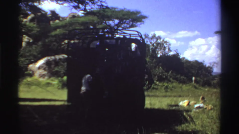 1969 Nigeria 8mm frame showing a dark truck parked beneath a tree as a person stands near its rear and items rest on the grass.