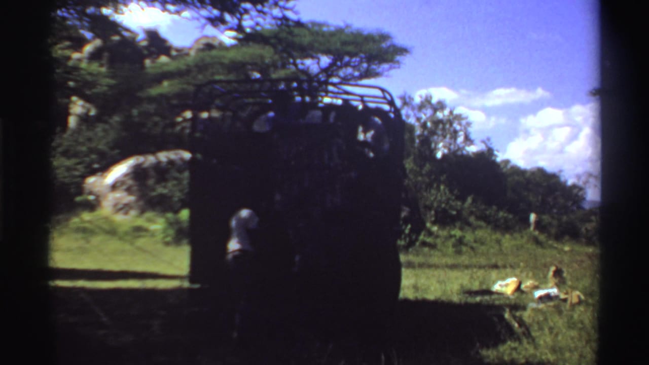 1969 Nigeria home-movie frame with a shaded truck, one visible person near the rear, and loose items on a sunny grassy area.