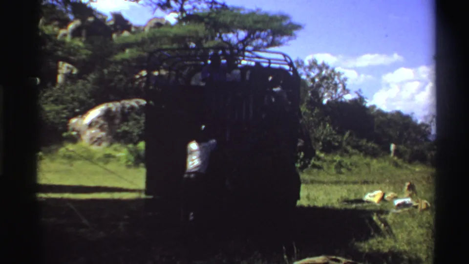1969 Nigeria 8mm frame showing people around a dark utility truck in grass, with trees, rocks, and a bright sky behind it.