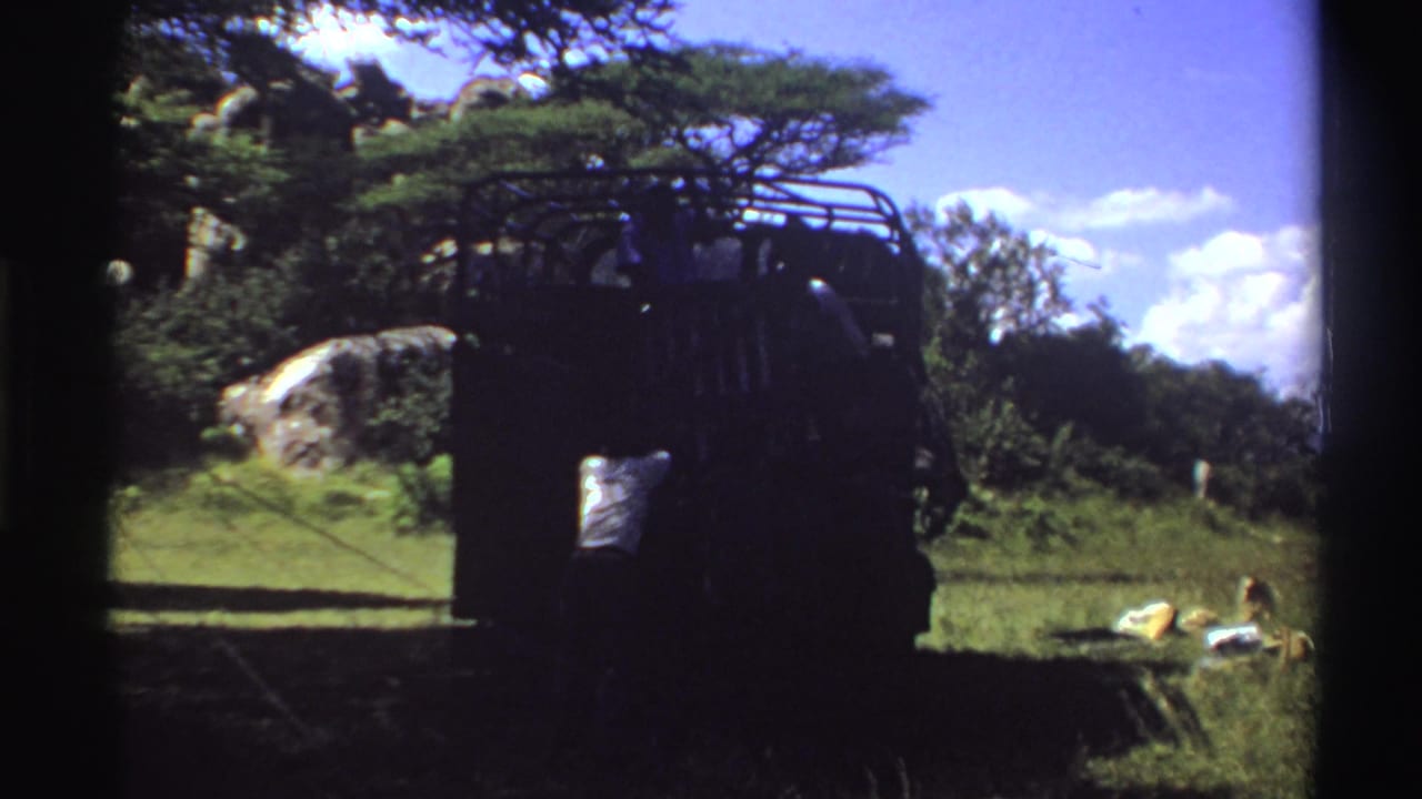 1969 Nigeria home-movie frame of a dark truck stopped in a grassy area, with people in shadow and small supplies nearby.