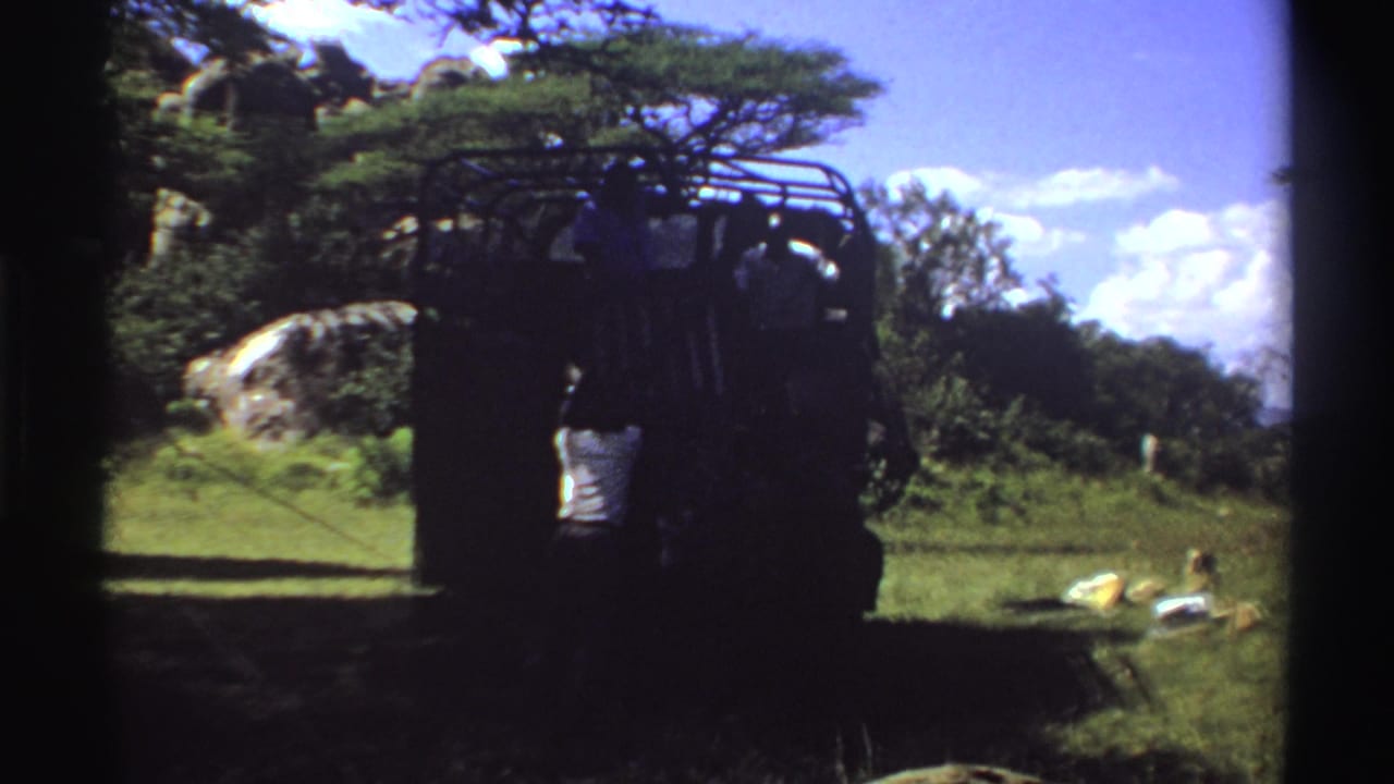 1969 Nigeria 8mm view of a utility truck in a rural field, framed by rocks, trees, grass, and strong midday contrast.