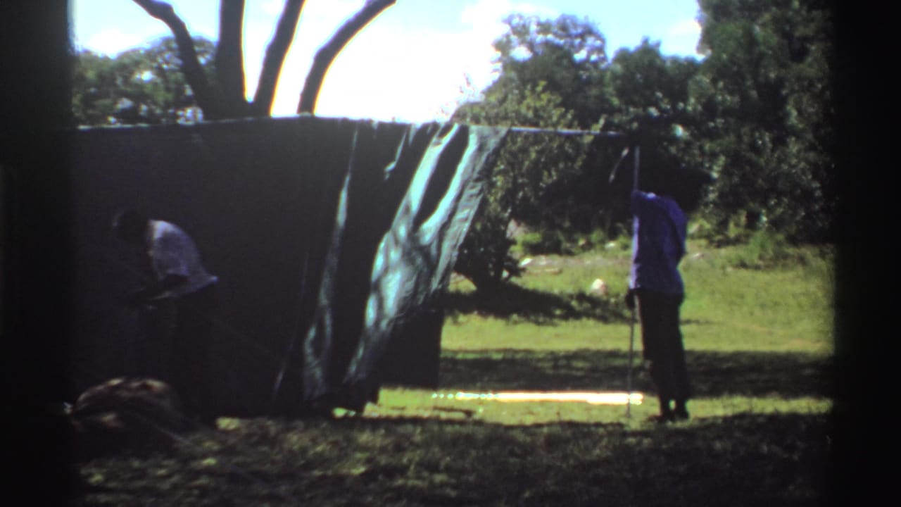 1969 Nigeria 8mm frame showing two people securing a large green tarp outdoors with trees and grass behind them.