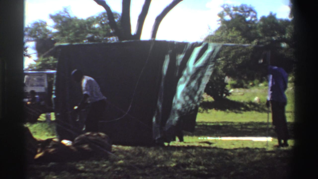 A 1969 Nigeria home-movie frame shows one person bending near the tarp base as another stands by a rope or pole.