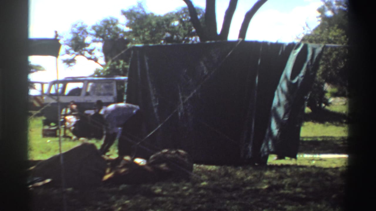 A white van is visible behind the green tarp while a person bends over ropes or gear in the grassy foreground.