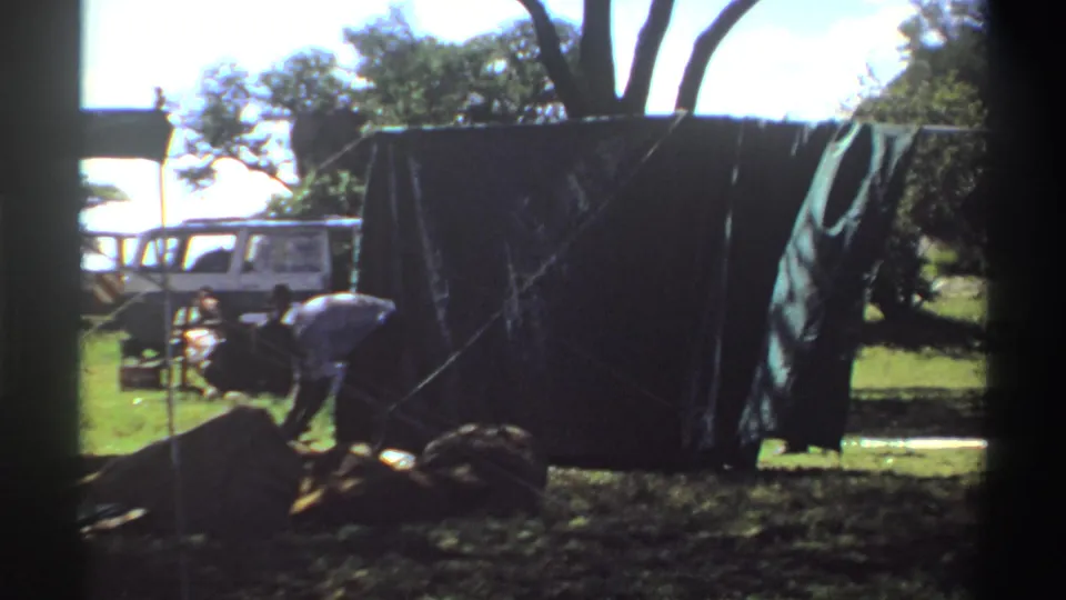 A white van is visible behind the green tarp while a person bends over ropes or gear in the grassy foreground.