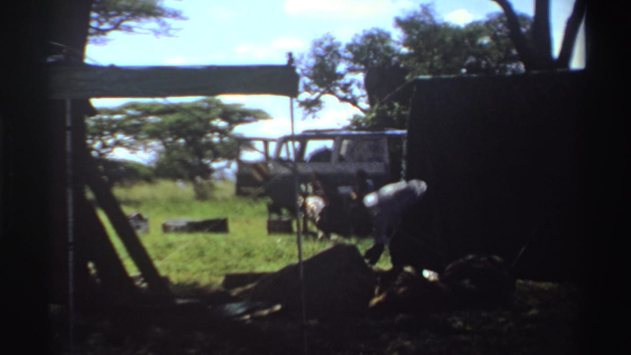 1969 Nigeria 8mm frame showing a man working near low furniture or equipment beside a parked white van in a grassy camp setting.