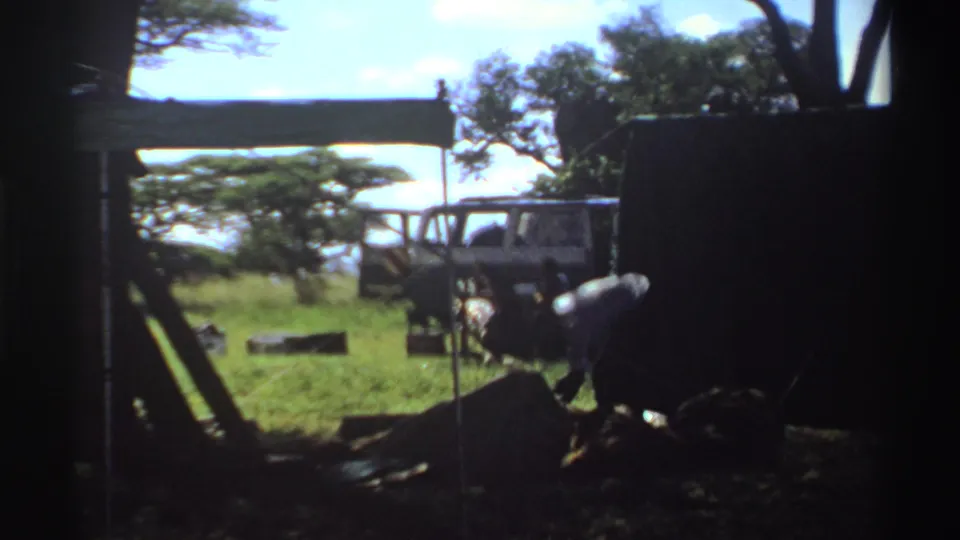 1969 Nigeria 8mm frame showing a man working near low furniture or equipment beside a parked white van in a grassy camp setting.
