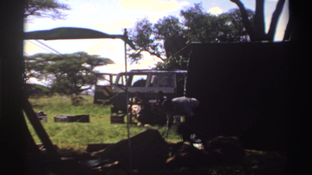 1969 Nigeria home-movie frame with a van under trees and people gathered near low camp furniture or equipment.