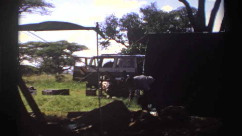 1969 Nigeria home-movie frame with a van under trees and people gathered near low camp furniture or equipment.
