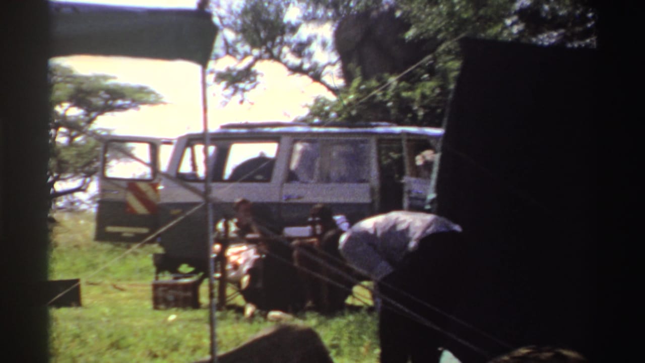 A closer 8mm view shows the parked van, open door, seated figures, and a man bending over camp furniture in Nigeria.
