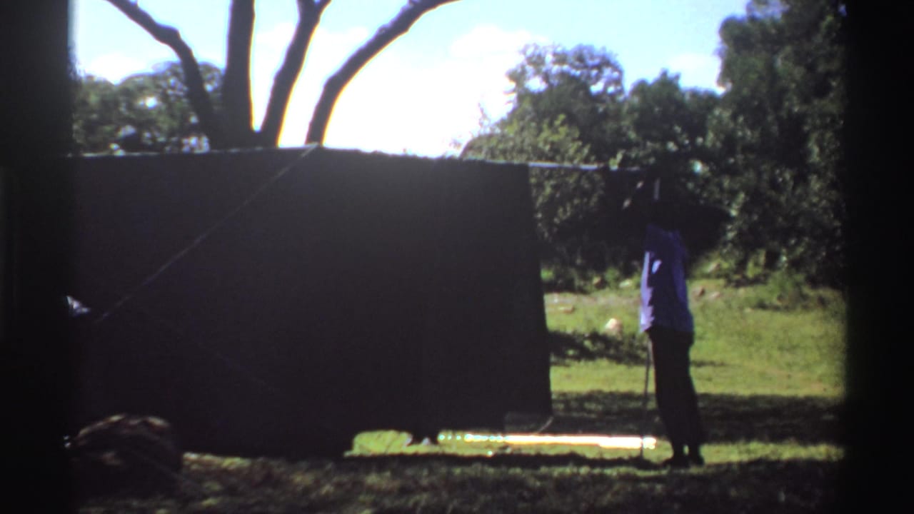 Person standing beside a dark tent in a grassy area with trees behind it.