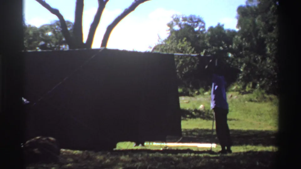1969 Nigeria 8mm frame showing a person beside a dark tent or screen in bright sun, with trees and grass in the background.
