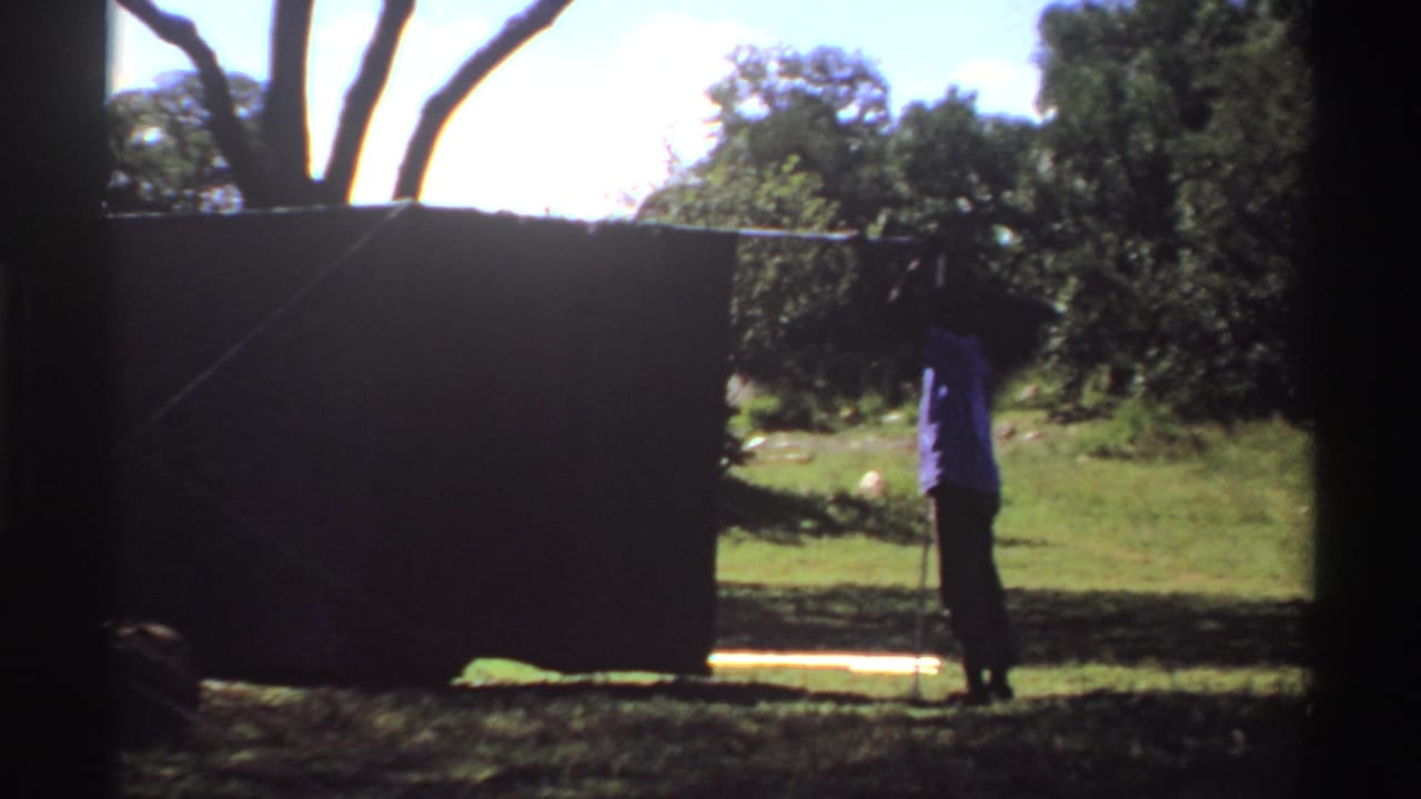 1969 Nigeria frame with a person standing near the right edge of a dark tent, surrounded by grass, trees, and strong daylight.