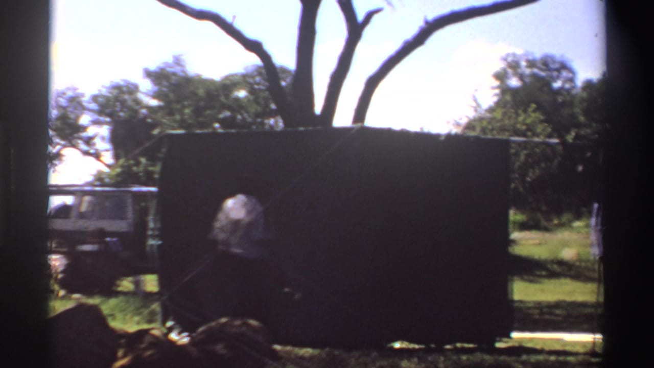 1969 Nigeria 8mm frame showing a dark tent in a grassy setting, with a white vehicle partly visible behind it and a person near the edge.