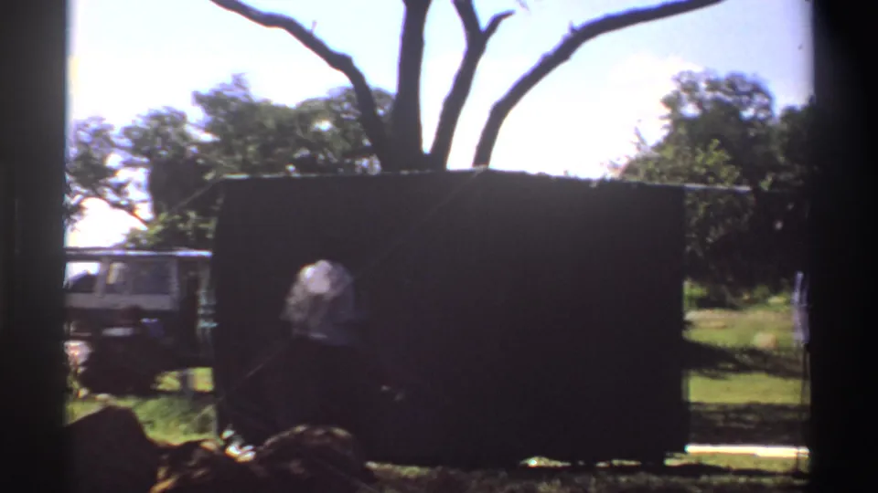 1969 Nigeria 8mm frame showing a dark tent in a grassy setting, with a white vehicle partly visible behind it and a person near the edge.