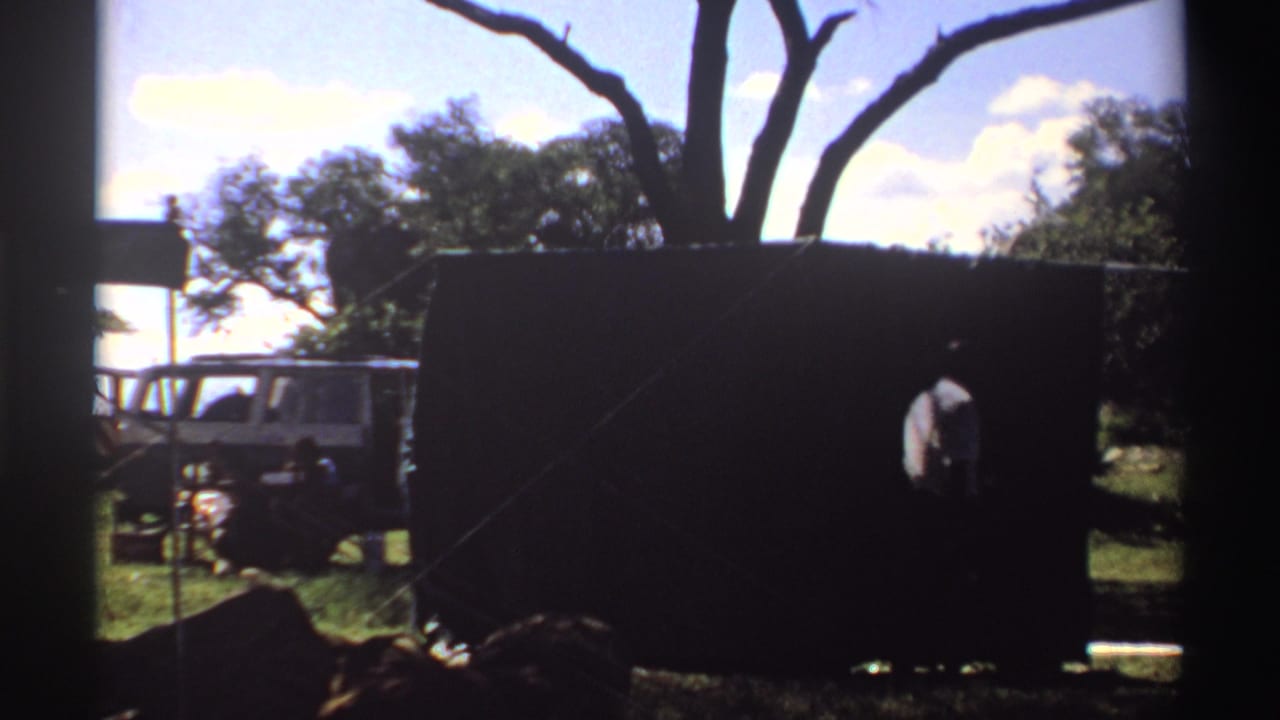 1969 Nigeria frame showing a person bending or working near a dark tent, with trees, grass, and a partly visible white vehicle in the background.