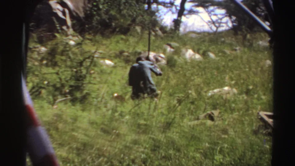 1969 Nigeria 8mm frame showing a man in tall savanna grass holding a long stick, with rocks and trees behind him.