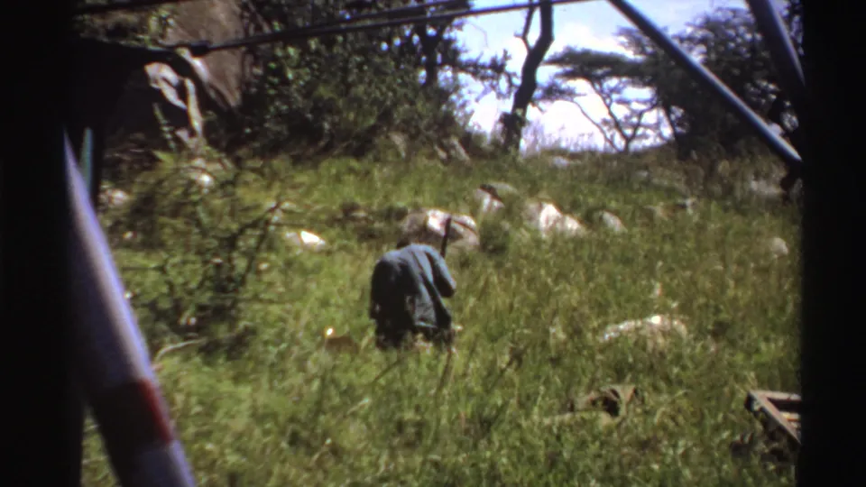 Late-1960s Nigeria frame showing a man digging or working in a grassy field, with vehicle structure at the edge of the view.