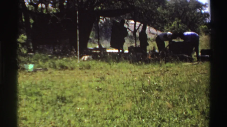 Distant 1969 Nigeria home-movie view of figures working in tree shade beyond tall grass.