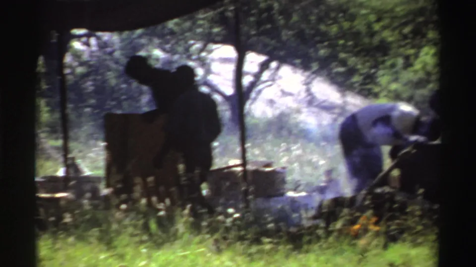 A shaded 1969 Nigeria frame shows people working beside containers as smoke drifts through the wooded camp area.