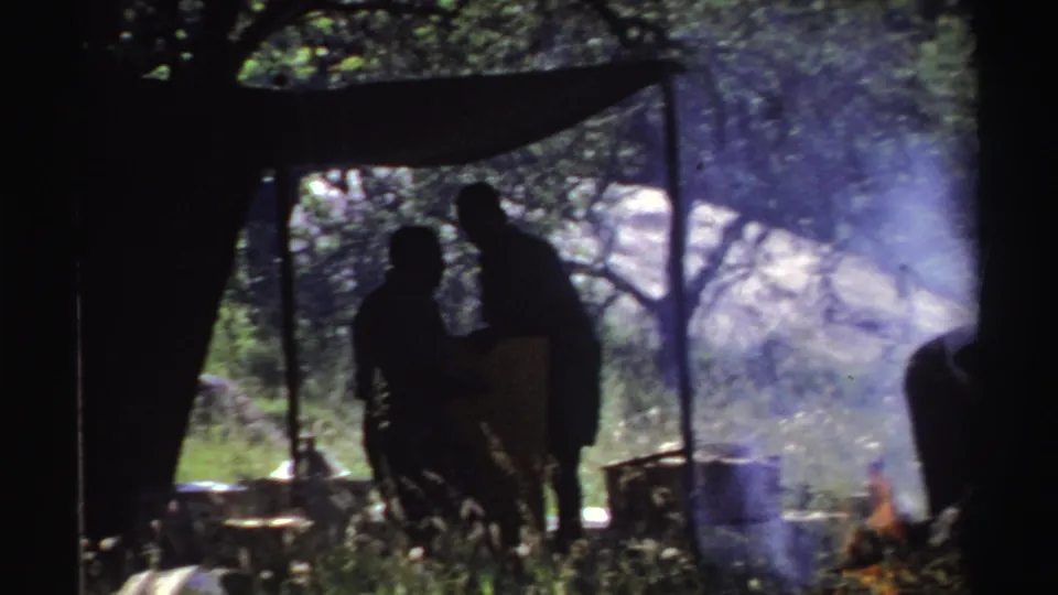 In the Nigeria 1969 clip, two figures stand near the tent as smoke spreads from a small fire in the clearing.