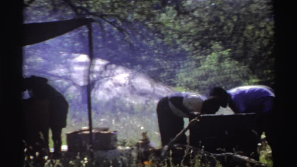 Nigeria 1969 8mm frame showing people gathered around an outdoor cooking pot, with smoke, trees, grass, and a rocky outcrop behind them.