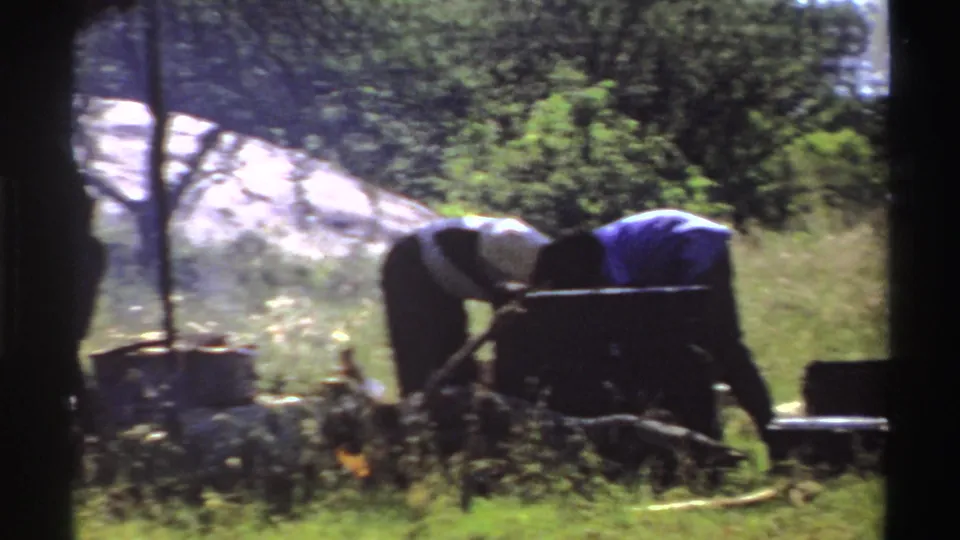 Nigeria 1969 8mm frame showing two people bent over a cooking pot near smoke, tall grass, trees, and a pale rocky outcrop.