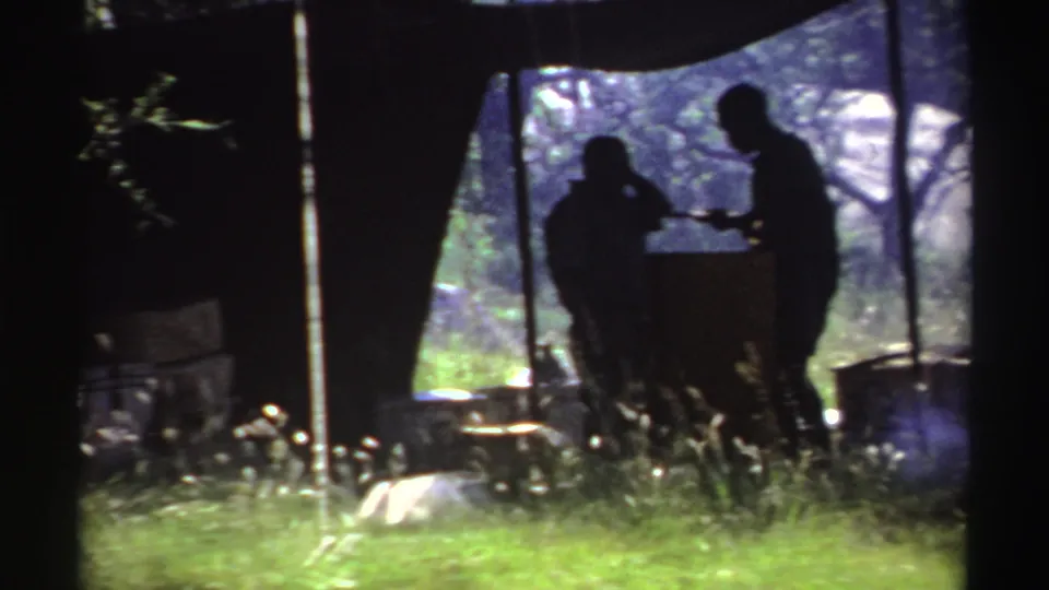 Nigeria 1969 8mm frame with silhouetted figures under a cloth shelter near an outdoor cooking setup, grass, trees, and drifting smoke.
