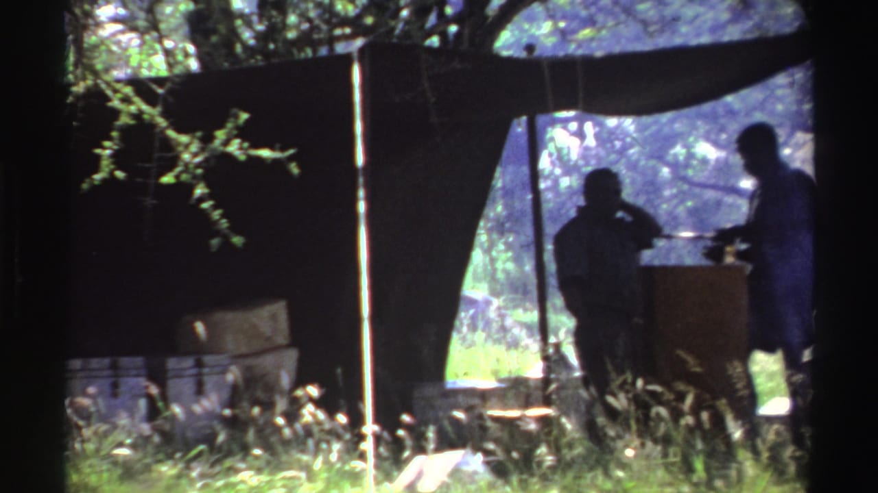 In 1969 Nigeria footage, two silhouetted men stand beneath a dark tarp shelter in a sunlit grassy outdoor area.