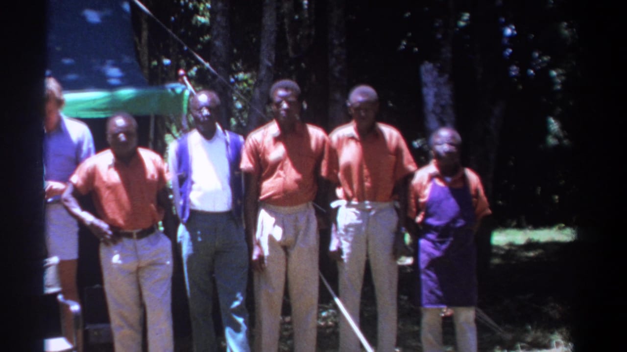 Men stand together outdoors near a canopy in bright sunlight.