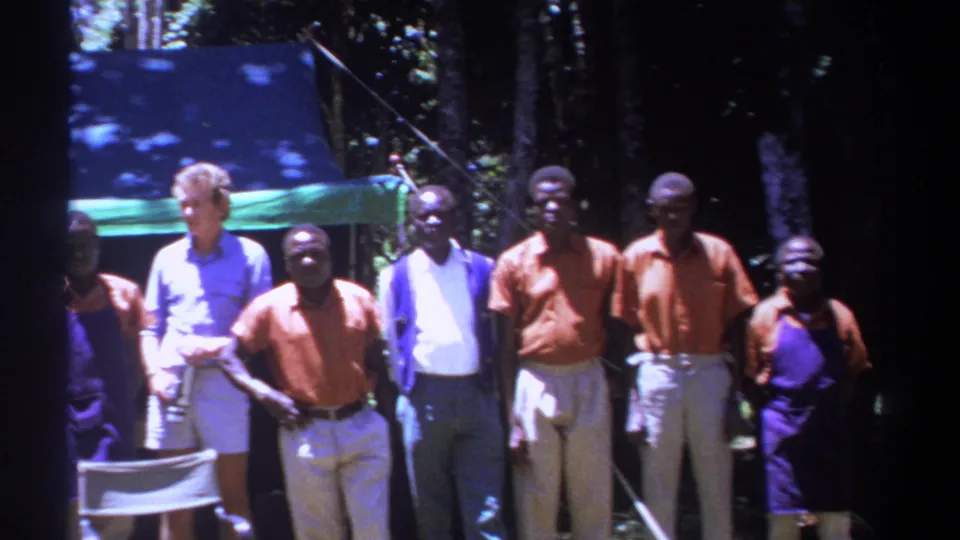 Kenya 1969 travel-film frame showing men gathered beside chairs and a canopy in a wooded area.
