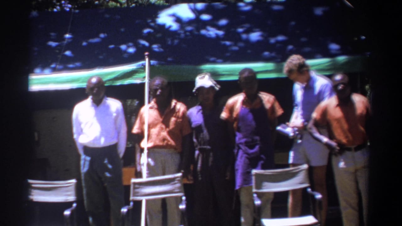 Men stand near director's chairs while a man in a blue shirt looks down at equipment on the right.