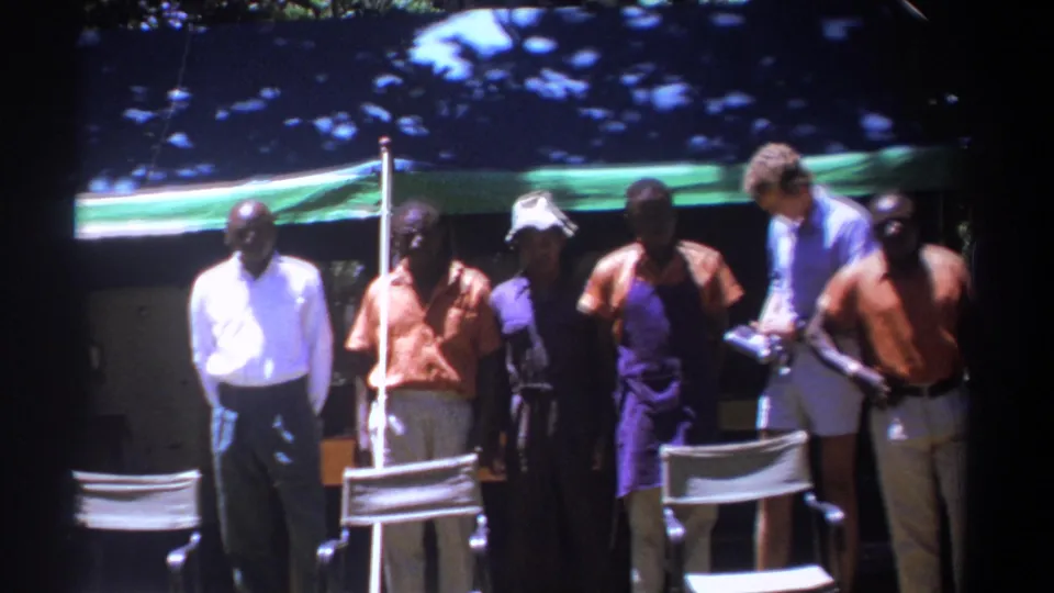 In Kenya 1969 footage, several adult men stand beneath an awning while a man in a blue shirt appears to handle equipment beside them.