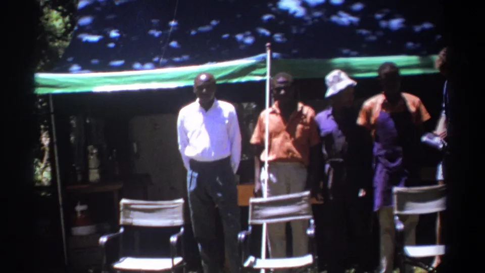 In Kenya 1969 footage, four adult men stand behind folding chairs under a green awning, with deep tree shade and bright sunlight across the scene.