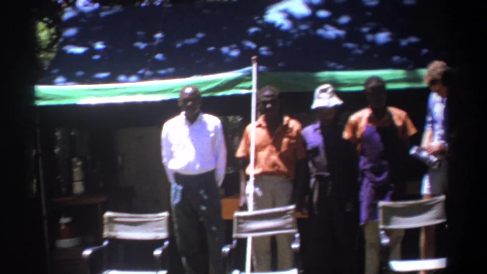 In Kenya 1969 footage, a row of adult men stands behind director's chairs beneath a green awning, with one figure partly cropped at the right edge.