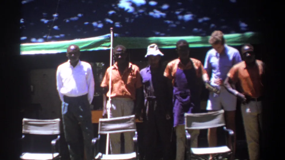 In Kenya 1969 footage, adult men in orange shirts and purple aprons stand outdoors near white chairs under a green canopy.