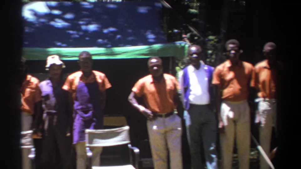 The 1969 Kenya home-movie frame shows men in orange shirts and purple aprons standing near a white chair in bright sun.