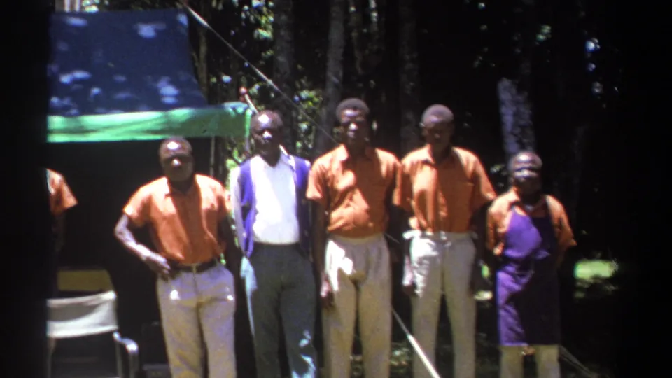 Adult men in orange shirts, light trousers, and purple aprons pose in a wooded Kenya setting on vintage 8mm footage.