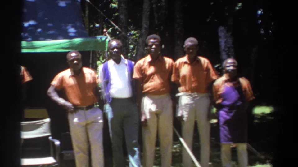 Adults in Kenya in 1969 stand for the camera near a green canopy, with orange shirts and a purple vest visible.