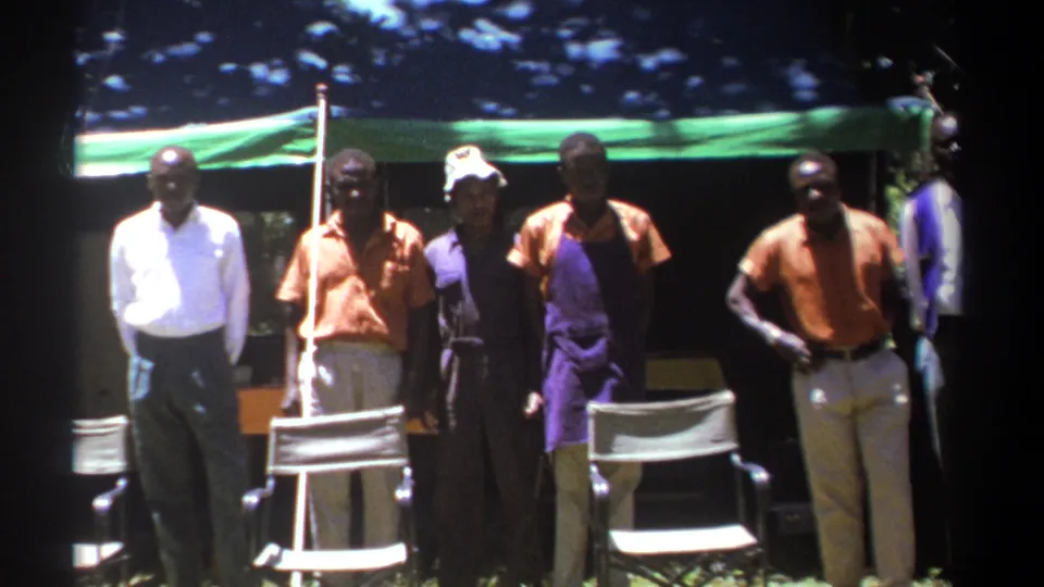 In a 1969 Kenya outdoor scene, adults pose beside folding chairs under a green canopy, framed by trees and harsh sunlight.