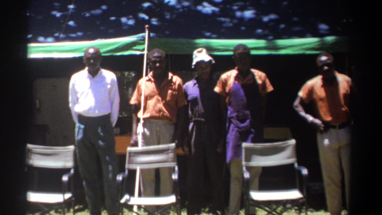 Group of men standing beneath a tarp beside folding chairs outdoors.